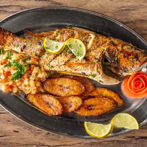 Whole grilled fish with lemon slices, fried plantains, and a vegetable medley on a silver platter.